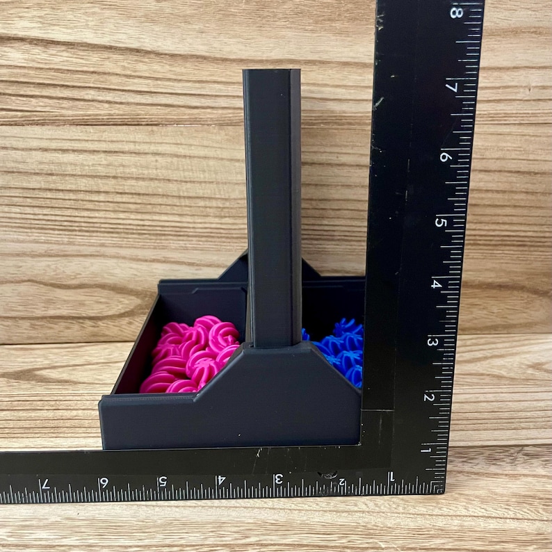 May include: A matte black plastic organiser with a central handle and two compartments, containing small pink and blue spiral-shaped pieces. A black L-shaped ruler is positioned against the side and base, showing a height of 20.32 cm.