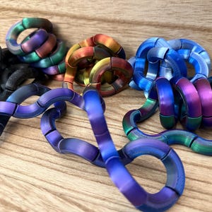 Puede incluir: Una colecci&oacute;n de juguetes sensoriales articulados hechos de segmentos curvos interconectados. Los juguetes presentan acabados met&aacute;licos en varios colores, incluyendo azul, morado, verde, naranja, amarillo iridiscente y negro mate, con superficies texturizadas.