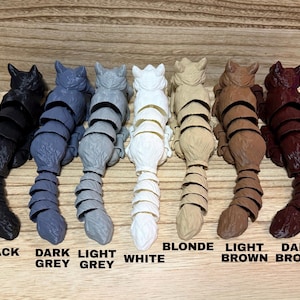 May include: Seven articulated cat figurines arranged in a row, each featuring a segmented tail and body. The colors from left to right are black, dark grey, light grey, white, blonde, light brown, and dark brown, with labels below each.