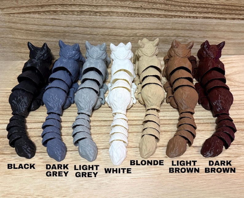 May include: Seven articulated cat figurines arranged in a row, each featuring a segmented tail and body. The colors from left to right are black, dark grey, light grey, white, blonde, light brown, and dark brown, with labels below each.
