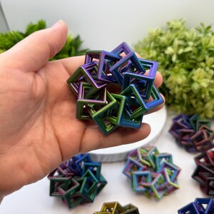 Pode incluir: Uma m&atilde;o segurando uma pequena escultura geom&eacute;trica iridescente composta por molduras quadradas interconectadas. A escultura exibe um gradiente de cores, incluindo azul, verde e roxo. Outras esculturas semelhantes s&atilde;o vis&iacute;veis no fundo.