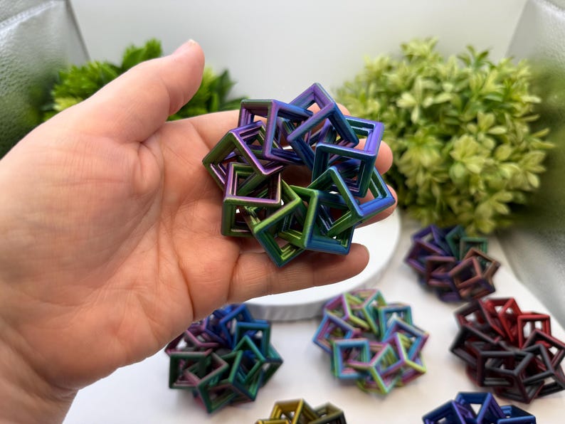 Pode incluir: Uma m&atilde;o segurando uma pequena escultura geom&eacute;trica iridescente composta por molduras quadradas interconectadas. A escultura exibe um gradiente de cores, incluindo azul, verde e roxo. Outras esculturas semelhantes s&atilde;o vis&iacute;veis no fundo.