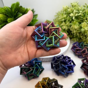 Pode incluir: Uma m&atilde;o segurando uma pequena escultura geom&eacute;trica iridescente composta por molduras quadradas interconectadas. V&aacute;rias esculturas semelhantes em v&aacute;rias cores, incluindo azul, roxo e vermelho, est&atilde;o dispostas sobre uma superf&iacute;cie branca, com folhagem verde ao fundo.