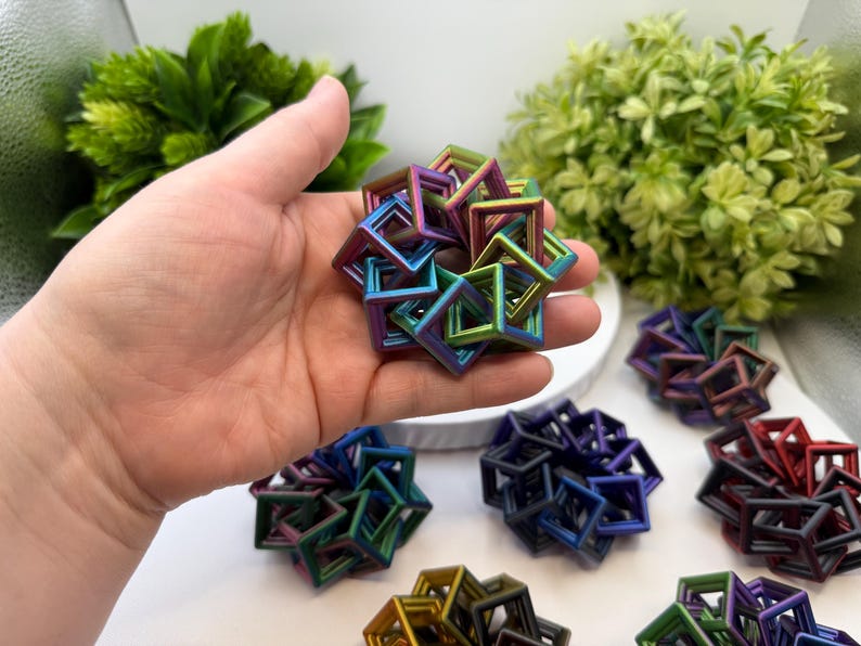 Pode incluir: Uma m&atilde;o segurando uma pequena escultura geom&eacute;trica iridescente composta por molduras quadradas interconectadas. V&aacute;rias esculturas semelhantes em v&aacute;rias cores, incluindo azul, roxo e vermelho, est&atilde;o dispostas sobre uma superf&iacute;cie branca, com folhagem verde ao fundo.