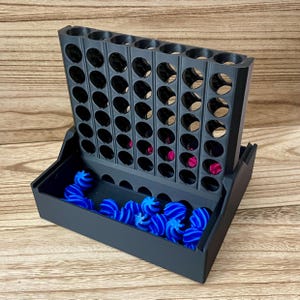 May include: A black plastic vertical board game with a grid of circular slots, featuring a base tray filled with small, bright blue spiral-shaped game pieces. A few magenta-coloured pieces are visible inside the lower slots of the grid structure.