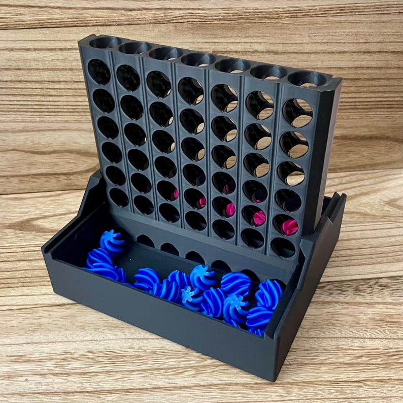 May include: A black plastic vertical board game with a grid of circular slots, featuring a base tray filled with small, bright blue spiral-shaped game pieces. A few magenta-coloured pieces are visible inside the lower slots of the grid structure.