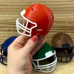 Puede incluir: Una colecci&oacute;n de cascos de f&uacute;tbol americano en miniatura de varios colores, incluyendo naranja, azul, verde y marr&oacute;n. Cada casco cuenta con una rejilla protectora y un dise&ntilde;o de carcasa redondeada. Estas r&eacute;plicas de pl&aacute;stico se muestran juntas.