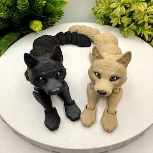 May include: Two articulated wolf figurines, one in solid black and one in light beige, featuring textured surfaces, pointed ears, and dark eyes. Each wolf has a segmented, flexible body and tail, positioned in a resting pose with front paws extended forward on a circular white surface.