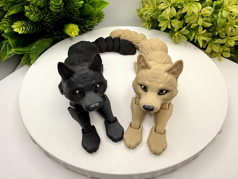 May include: Two articulated wolf figurines, one in solid black and one in light beige, featuring textured surfaces, pointed ears, and dark eyes. Each wolf has a segmented, flexible body and tail, positioned in a resting pose with front paws extended forward on a circular white surface.