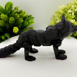 May include: A matte black articulated fox figurine featuring a segmented body design that allows for movement. The fox has pointed ears, a textured coat, and a long tail, standing on four legs with visible joints at the shoulders and hips.