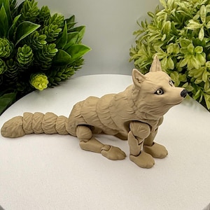 May include: A tan-colored articulated wolf figurine featuring a segmented body and tail, pointed ears, and dark eyes. The figure is posed in a standing position with visible joints at the shoulders, hips, and along the length of the tail.
