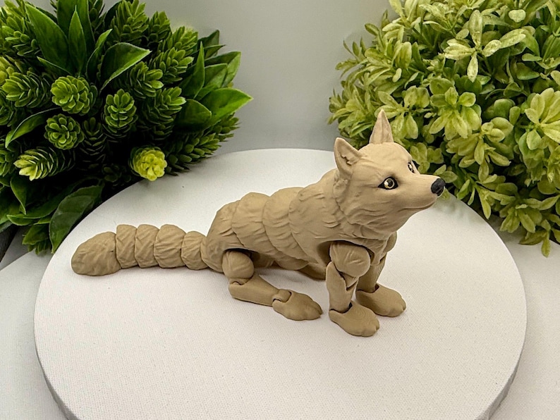 May include: A tan-colored articulated wolf figurine featuring a segmented body and tail, pointed ears, and dark eyes. The figure is posed in a standing position with visible joints at the shoulders, hips, and along the length of the tail.