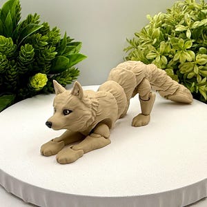 May include: A tan articulated wolf figurine in a crouching position. The figure features a textured coat, pointed ears, and dark eyes. The body is composed of multiple segments connected by visible joints, allowing for poseability of the limbs and tail.