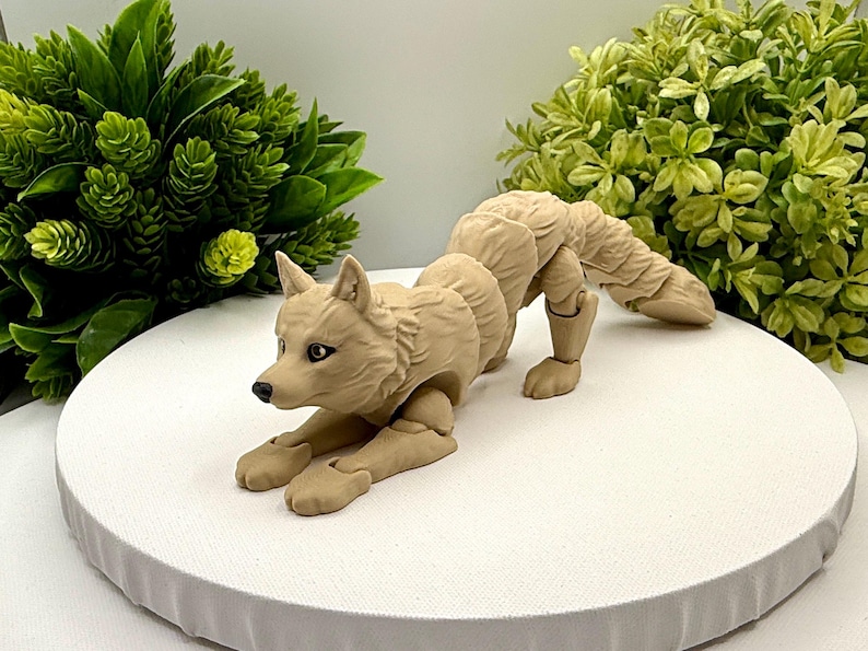 May include: A tan articulated wolf figurine in a crouching position. The figure features a textured coat, pointed ears, and dark eyes. The body is composed of multiple segments connected by visible joints, allowing for poseability of the limbs and tail.
