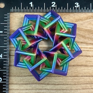 Pode incluir: Uma escultura geom&eacute;trica multicolorida composta por formas quadradas interconectadas. A escultura exibe um gradiente de cores, incluindo azul, verde e roxo. O objeto &eacute; colocado sobre uma superf&iacute;cie de madeira ao lado de uma r&eacute;gua.