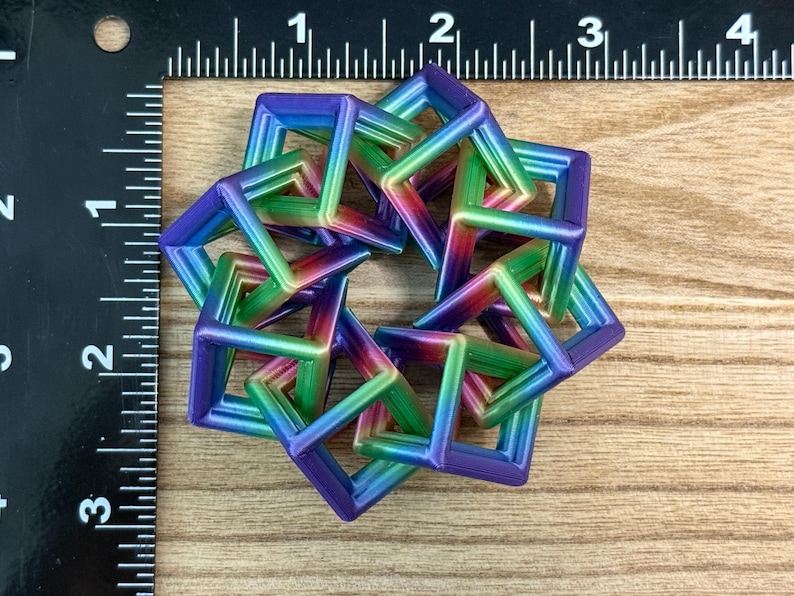 Pode incluir: Uma escultura geom&eacute;trica multicolorida composta por formas quadradas interconectadas. A escultura exibe um gradiente de cores, incluindo azul, verde e roxo. O objeto &eacute; colocado sobre uma superf&iacute;cie de madeira ao lado de uma r&eacute;gua.