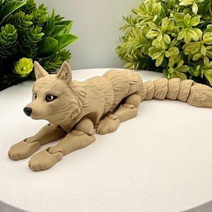 May include: A tan-colored articulated wolf figurine with a segmented body and tail. The figure features molded fur texture, dark eyes, and a black nose. The design allows for flexible movement of the limbs and tail, showcasing a realistic canine posture in a lying position with front paws extended forward.