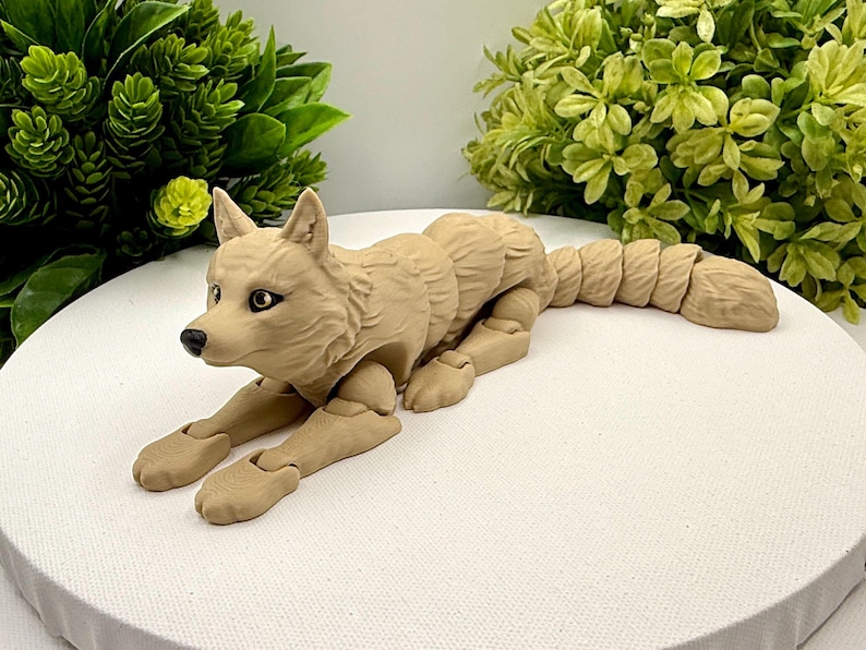May include: A tan-colored articulated wolf figurine with a segmented body and tail. The figure features molded fur texture, dark eyes, and a black nose. The design allows for flexible movement of the limbs and tail, showcasing a realistic canine posture in a lying position with front paws extended forward.