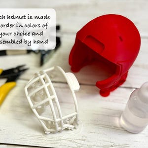 Puede incluir: Una carcasa de casco de color rojo brillante y una m&aacute;scara facial blanca, acompa&ntilde;adas de unos alicates con mangos amarillos y un peque&ntilde;o frasco de adhesivo transparente. El texto dice: Each helmet is made to order in colors of your choice and assembled by hand.