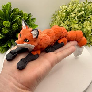 May include: An articulated fox figurine featuring a bright orange body with textured fur segments, black legs and paws, and white accents on the ears and face. The fox has a segmented, flexible tail and dark eyes, designed with a multi-jointed structure.