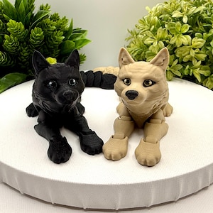 May include: Two articulated wolf figurines in a lying position, one solid black and one light tan. Both figures feature detailed fur textures, pointed ears, and dark eyes. The wolves are positioned side-by-side, showcasing their jointed limbs and realistic facial features in a compact, sculptural form.