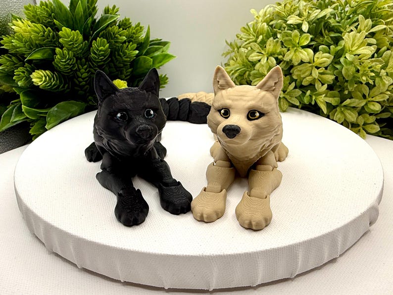 May include: Two articulated wolf figurines in a lying position, one solid black and one light tan. Both figures feature detailed fur textures, pointed ears, and dark eyes. The wolves are positioned side-by-side, showcasing their jointed limbs and realistic facial features in a compact, sculptural form.