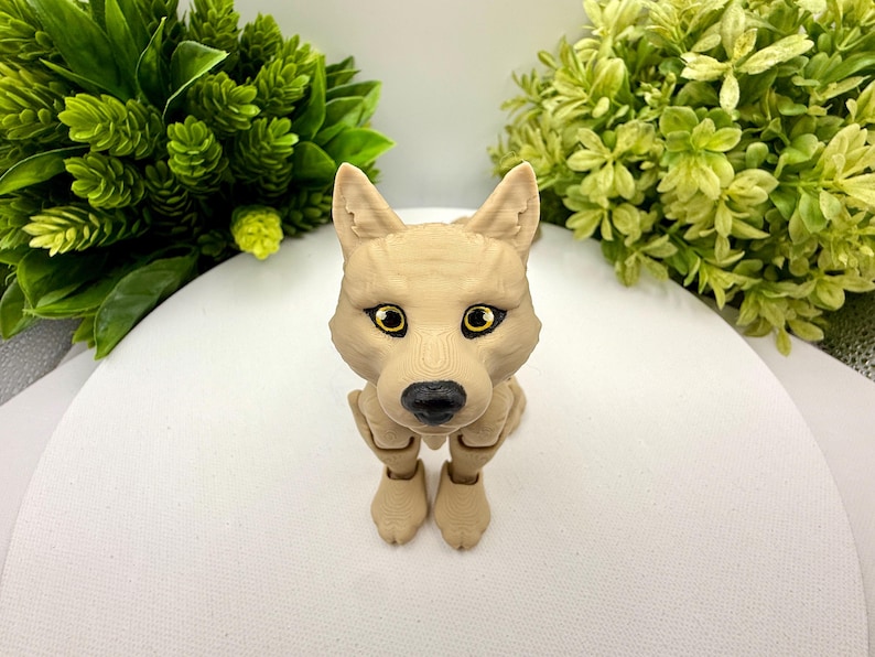 May include: A tan-colored articulated wolf figurine with a smooth, matte finish. The figure features pointed ears, a black nose, and large, expressive eyes with yellow irises and black pupils. The body is sculpted with visible texture lines.