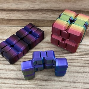 May include: Three colorful fidget toys in shades of purple, blue, and rainbow. The toys are made of connected blocks that can be manipulated. The background is a light brown wooden surface.