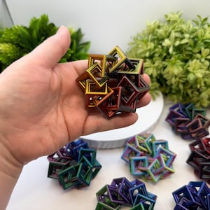 Pode incluir: Uma m&atilde;o segurando uma escultura geom&eacute;trica multicolorida composta por quadrados interconectados. A escultura tem um acabamento met&aacute;lico com tons de ouro, vermelho e verde. V&aacute;rias esculturas semelhantes em v&aacute;rias cores est&atilde;o dispostas em uma superf&iacute;cie branca.