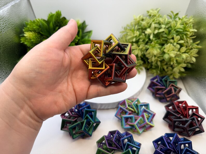Pode incluir: Uma m&atilde;o segurando uma escultura geom&eacute;trica multicolorida composta por quadrados interconectados. A escultura tem um acabamento met&aacute;lico com tons de ouro, vermelho e verde. V&aacute;rias esculturas semelhantes em v&aacute;rias cores est&atilde;o dispostas em uma superf&iacute;cie branca.