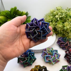 Pode incluir: Uma m&atilde;o segurando uma escultura geom&eacute;trica multicolorida composta por cubos interconectados. V&aacute;rias esculturas semelhantes em v&aacute;rias cores, incluindo azul, verde e vermelho, est&atilde;o dispostas sobre uma superf&iacute;cie branca. As esculturas t&ecirc;m um brilho met&aacute;lico.