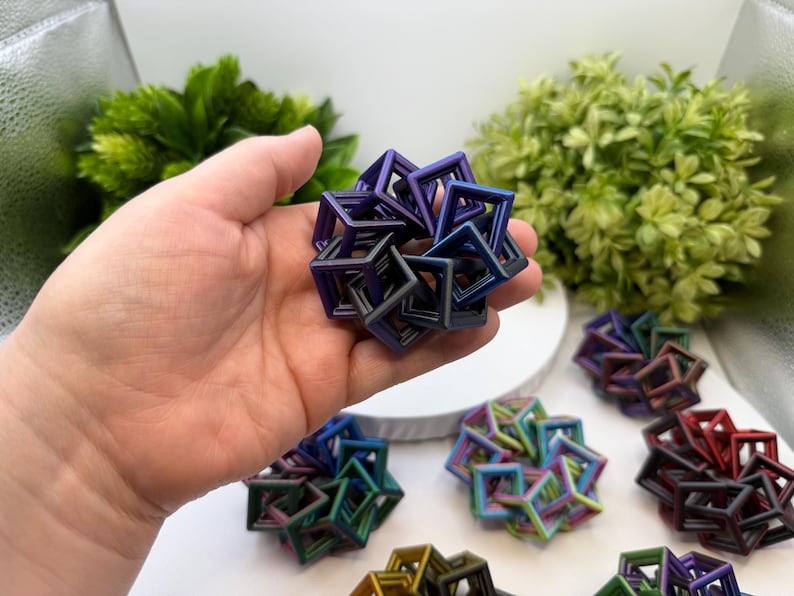 Pode incluir: Uma m&atilde;o segurando uma escultura geom&eacute;trica multicolorida composta por cubos interconectados. V&aacute;rias esculturas semelhantes em v&aacute;rias cores, incluindo azul, verde e vermelho, est&atilde;o dispostas sobre uma superf&iacute;cie branca. As esculturas t&ecirc;m um brilho met&aacute;lico.