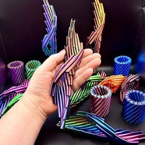 May include: A collection of 3D printed, articulated, flexible dragon-like sculptures and cylindrical containers in a vibrant rainbow gradient of metallic colours including blue, green, purple, orange, and pink, displayed in a dark studio setting.