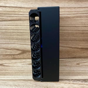 May include: A matte black rectangular organiser featuring a vertical row of seven circular storage compartments. Each compartment contains a coiled metal spring mechanism. A small blue circular accent is visible on the side of the black frame.