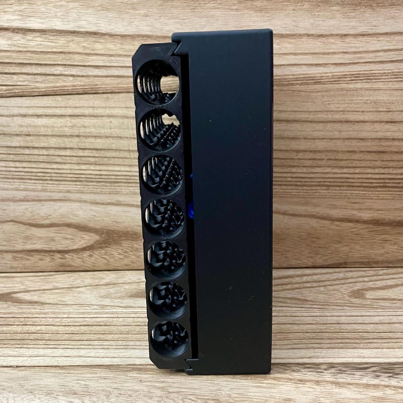 May include: A matte black rectangular organiser featuring a vertical row of seven circular storage compartments. Each compartment contains a coiled metal spring mechanism. A small blue circular accent is visible on the side of the black frame.