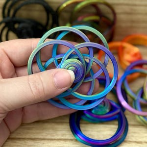 May include: A circular fidget spinner featuring an intricate, multi-layered geometric design with overlapping rings. The object displays a vibrant, iridescent rainbow gradient finish, shifting between shades of blue, purple, and green across its surface.