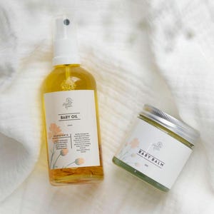 May include: A set of baby care products, a clear glass bottle of baby oil with a white spray top and a green jar of baby balm. Both products have a white label with the text "Little Flower" and the product name. The products are on a white textured surface.