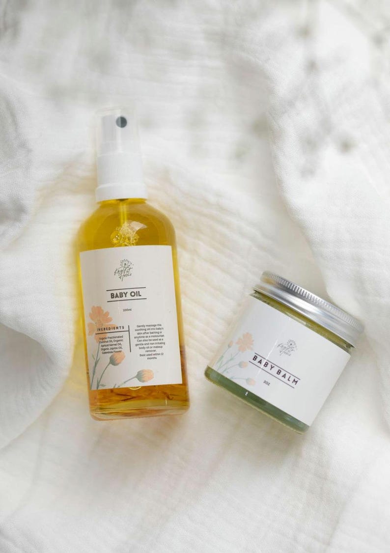 May include: A set of baby care products, a clear glass bottle of baby oil with a white spray top and a green jar of baby balm. Both products have a white label with the text "Little Flower" and the product name. The products are on a white textured surface.