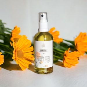 May include: A clear glass bottle with a white spray top filled with a yellow liquid. The bottle is labeled "Baby Oil" and lists ingredients including organic calendula oil. The bottle is sitting on a white surface with yellow flowers in the background.
