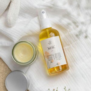 May include: A clear glass jar with a yellow balm inside, a silver lidded jar, and a clear glass bottle with a yellow liquid and a white label that says "Baby Oil" with a floral design. The items are on a white fabric background.