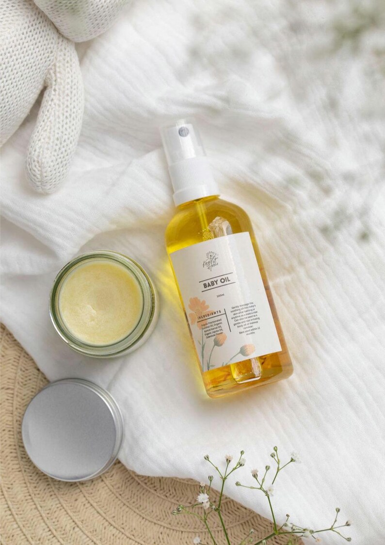 May include: A clear glass jar with a yellow balm inside, a silver lidded jar, and a clear glass bottle with a yellow liquid and a white label that says "Baby Oil" with a floral design. The items are on a white fabric background.
