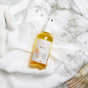 May include: A clear glass bottle of baby oil with a white spray nozzle and a white label that says "Baby Oil" with a floral design. The bottle is sitting on a white fabric surface.