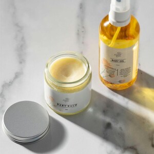 May include: A set of two baby care products, a white jar of baby balm and a clear glass bottle of baby oil. Both products have a white label with the text "Poplar & Vine" and the product name. The baby balm jar has a silver lid.
