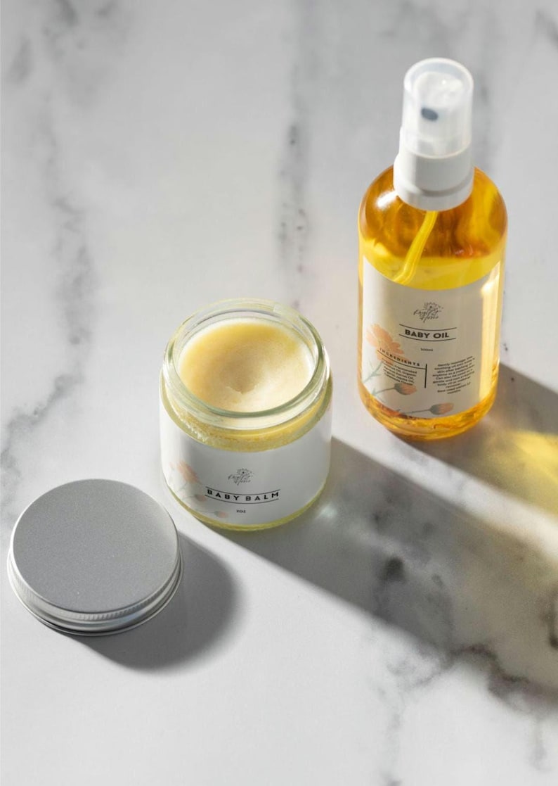 May include: A set of two baby care products, a white jar of baby balm and a clear glass bottle of baby oil. Both products have a white label with the text "Poplar & Vine" and the product name. The baby balm jar has a silver lid.