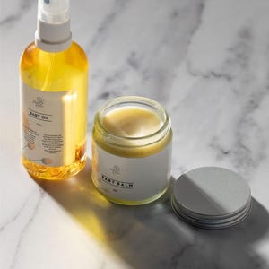 May include: Two glass bottles of baby care products on a white marble surface. The bottle on the left is a clear amber glass bottle with a white spray top and a label that reads "Baby Oil". The bottle on the right is a clear glass jar with a white label that reads "Baby Balm". The jar has a silver lid.