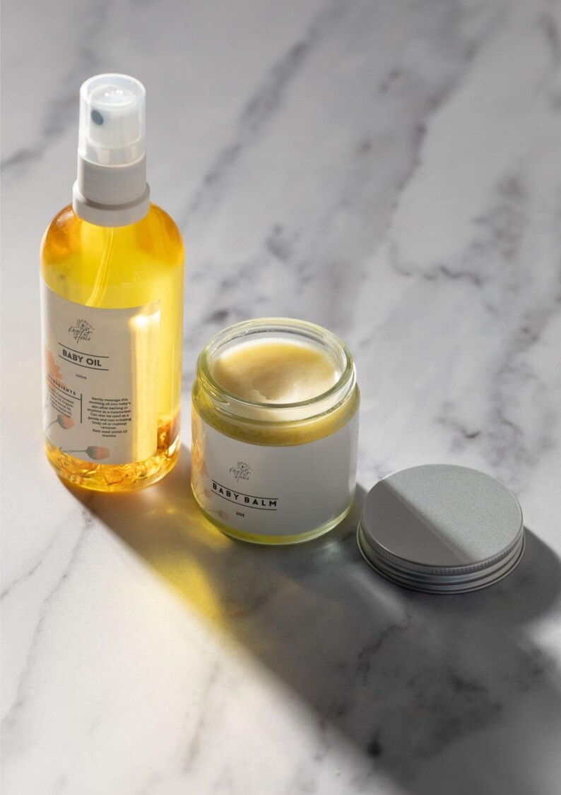 May include: Two glass bottles of baby care products on a white marble surface. The bottle on the left is a clear amber glass bottle with a white spray top and a label that reads "Baby Oil". The bottle on the right is a clear glass jar with a white label that reads "Baby Balm". The jar has a silver lid.