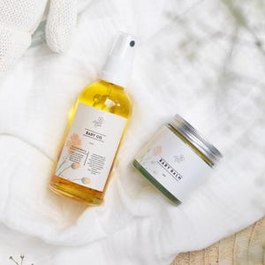 May include: A set of baby care products on a white fabric background. The set includes a bottle of baby oil with a white pump and a jar of baby balm with a silver lid. The labels on both products feature a floral design and the text "Baby Oil" and "Baby Balm".