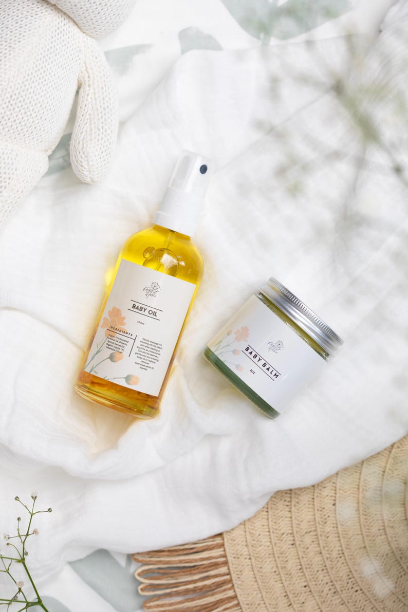 May include: A set of baby care products on a white fabric background. The set includes a bottle of baby oil with a white pump and a jar of baby balm with a silver lid. The labels on both products feature a floral design and the text "Baby Oil" and "Baby Balm".