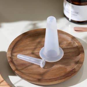 May include: A clear, cone-shaped silicone product with a wide base and a smaller, cylindrical piece, displayed on a round wooden tray. The items are likely related to skincare or beauty routines.