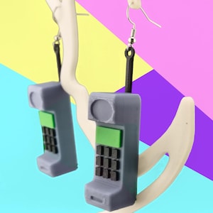 Könnte beinhalten: Ein Paar Hängeohrringe in Form von grauen Miniatur-Retro-Mobiltelefonen. Jedes Telefon verfügt über einen hellgrünen Bildschirmbereich und eine schwarze Tastatur mit neun Tasten. Die Hörer sind mit schwarzen Gliedern an silberfarbenen Metallhaken befestigt.